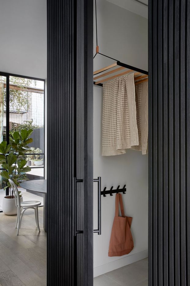 A contemporary interior view of a compact entryway or mudroom storage nook, framed by black vertical fluted timber-panelled doors. The nook has pale walls, a hanging rail with woven textiles, black hooks, and a terracotta tote bag. Through the adjacent opening is a bright dining area with a round dark table, chairs, large glazing, and indoor plants. The space uses a restrained neutral palette with strong black framing and warm terracotta accents.