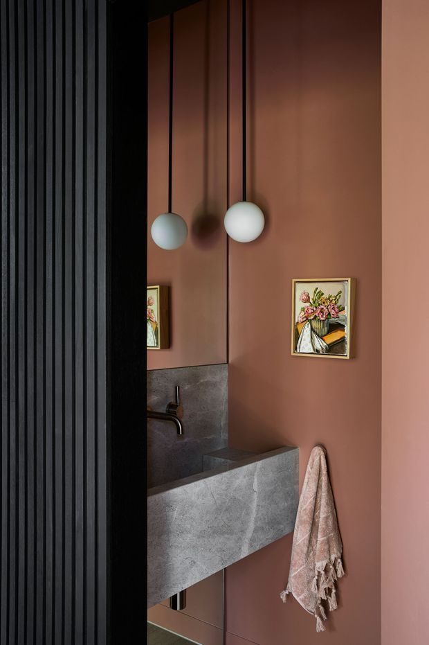 A narrow, moody bathroom interior viewed through a black vertical-slat partition. The room features dusty terracotta-pink walls, a large mirror, a floating grey stone or concrete vanity with an integrated basin, dark wall-mounted tapware, two hanging spherical pendant lights, and a framed floral artwork. A textured pink towel hangs beside the vanity.