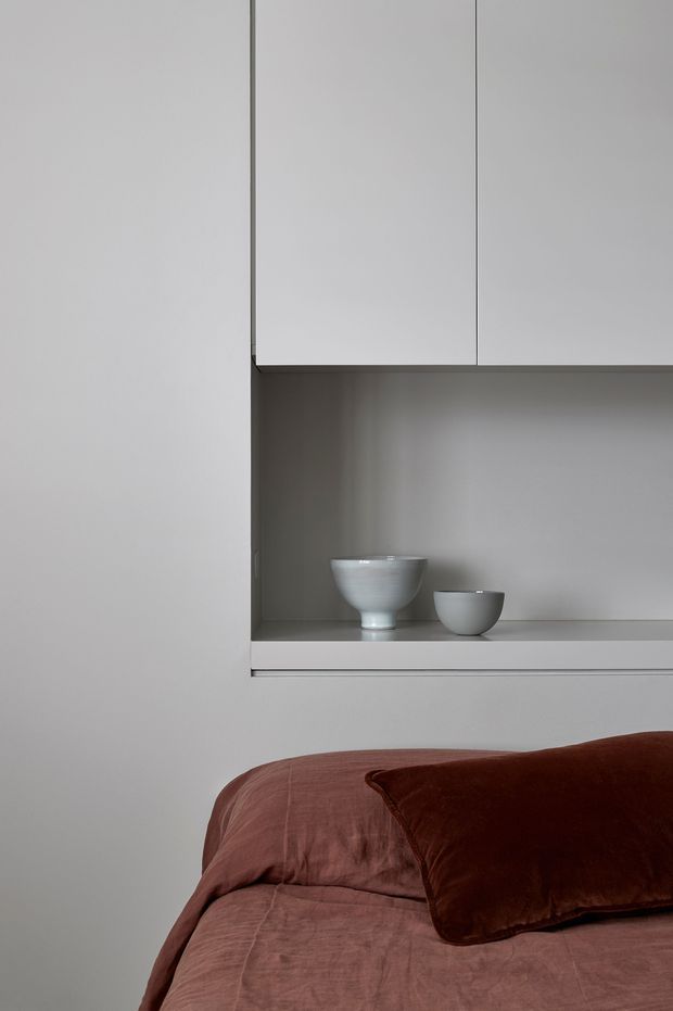 A tightly framed minimalist bedroom detail showing a bed with muted dusty-rose bedding and a dark rust-brown velvet cushion in the foreground. Above the bed is a recessed built-in wall niche with pale grey cabinetry and a shelf displaying two ceramic bowls. The palette is subdued and monochromatic, with clean joinery and smooth painted surfaces.
