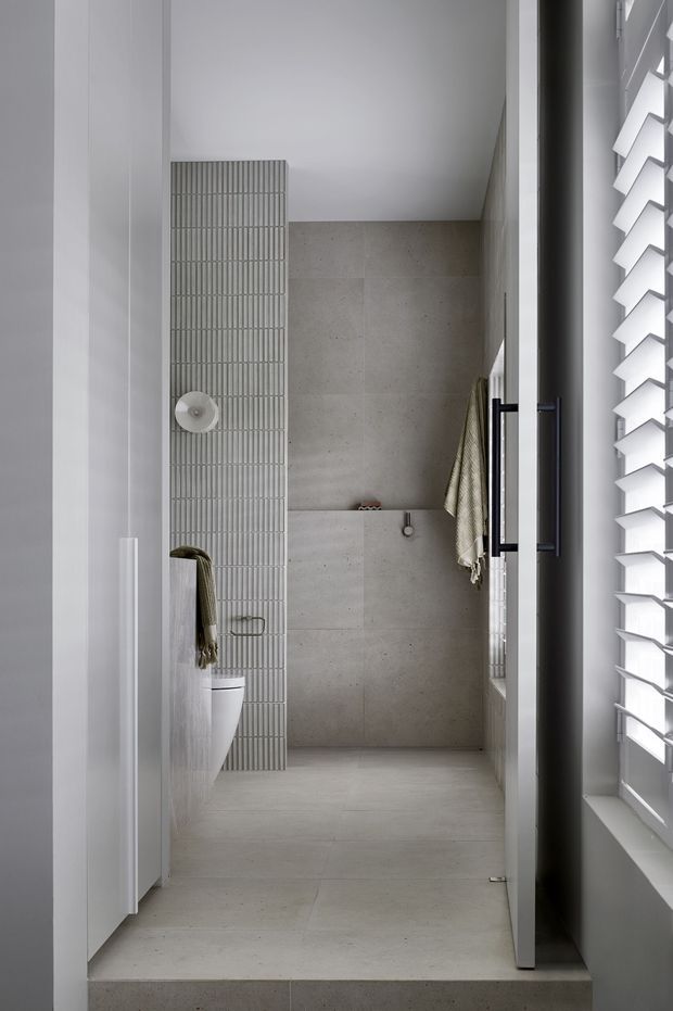A compact contemporary bathroom viewed from the doorway, featuring pale large-format stone-look wall and floor tiles, a vertical ribbed feature-tile panel, wall-hung toilet, frameless glass shower screen, dark hardware, and a white louvred window providing filtered daylight.