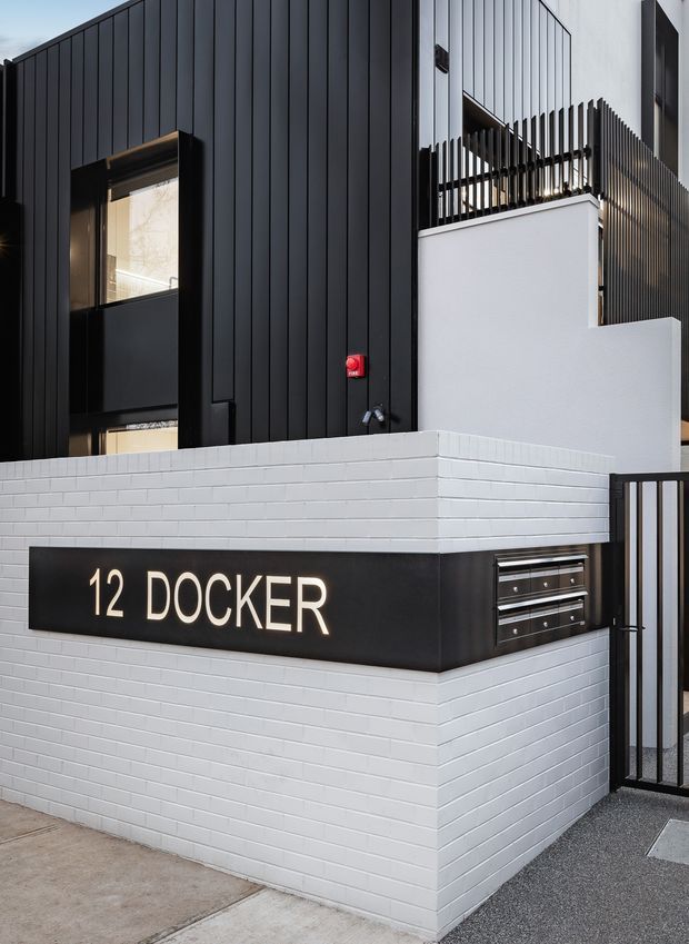 Street-level exterior view of a contemporary multi-storey residential building entrance. The facade combines dark vertical metal cladding with white-painted brickwork, a black metal gate, balcony screening, and a prominent black entry sign reading “12 DOCKER.”