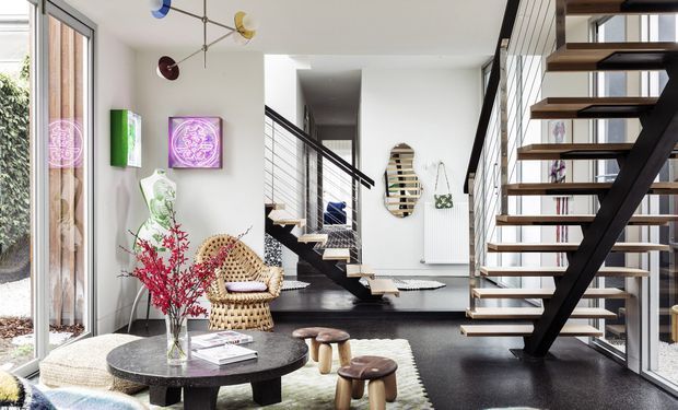 Bright contemporary open-plan interior centred on a sculptural open-riser staircase with dark timber or metal framing and cable balustrades. The space includes a lounge or entry area with a round coffee table, woven lounge chair, stools, artwork, decorative objects, and extensive floor-to-ceiling glazing. A second staircase and upper-level circulation are visible in the background. The palette is predominantly white, black, natural timber, and muted accent colours, with a refined modern aesthetic.