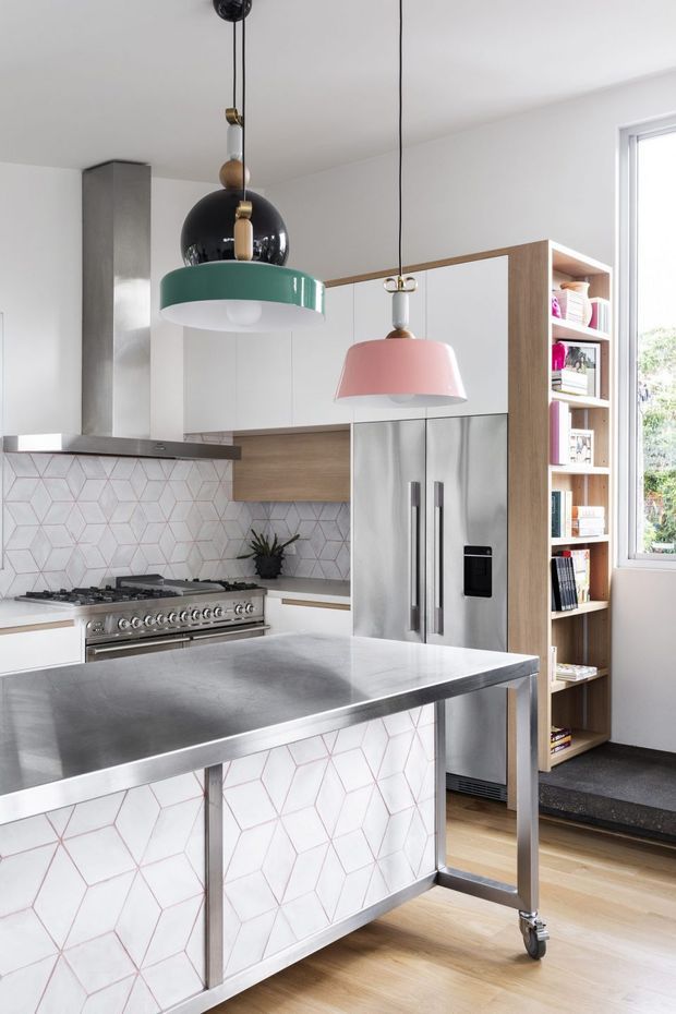 Bright contemporary kitchen featuring a large stainless-steel island on caster wheels, a stainless rangehood and commercial-style cooker, white cabinetry with warm timber accents, geometric white-and-pink tiled splashback, integrated stainless refrigerator, open timber shelving, and distinctive green, black, and pink pendant lights. A large window provides natural light.