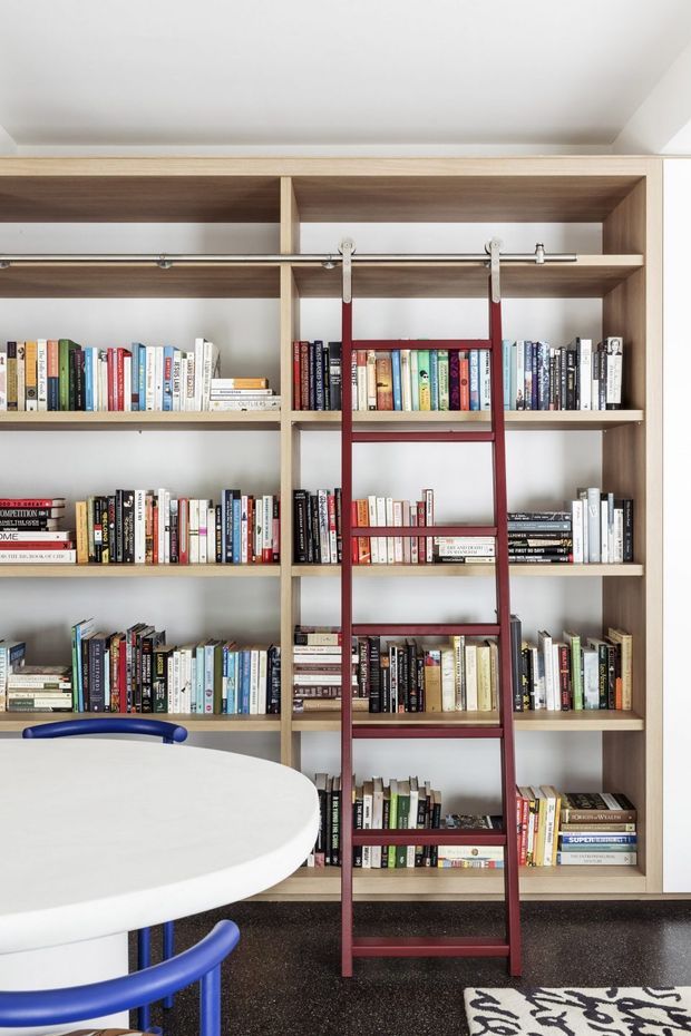 Bright interior featuring a full-height built-in timber bookshelf filled with books, with a distinctive dark red rolling library ladder mounted on a horizontal rail. A white oval table and vivid blue chairs occupy the foreground, while patterned carpeting and a monochrome rug add contrast.