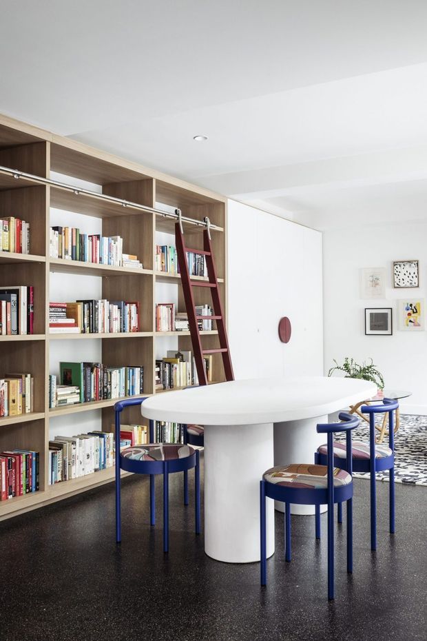 Bright contemporary open-plan dining and library space featuring a full-height timber built-in bookshelf with rolling ladder, a large white oval dining table, and sculptural blue dining chairs. White walls and ceiling, dark speckled flooring, framed artwork, and a small plant create a crisp, playful, design-focused interior.