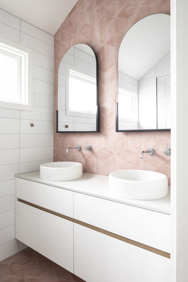 Bright contemporary bathroom featuring a double white vessel-basin vanity, wall-mounted chrome tapware, white tiled walls, and a pink blush geometric feature-tile wall that continues onto the floor. Two arched black-framed mirrors sit above the vanity, with natural light entering through small windows.