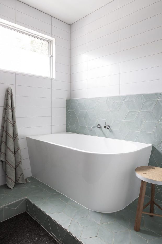 Bright contemporary bathroom featuring a freestanding white bathtub set against white horizontal wall tiles and a pale blue-green geometric hexagonal tile splashback. Matching hexagonal floor tiles continue onto a raised platform, with a high horizontal window, wall-mounted bath fixtures, and a small wooden stool.