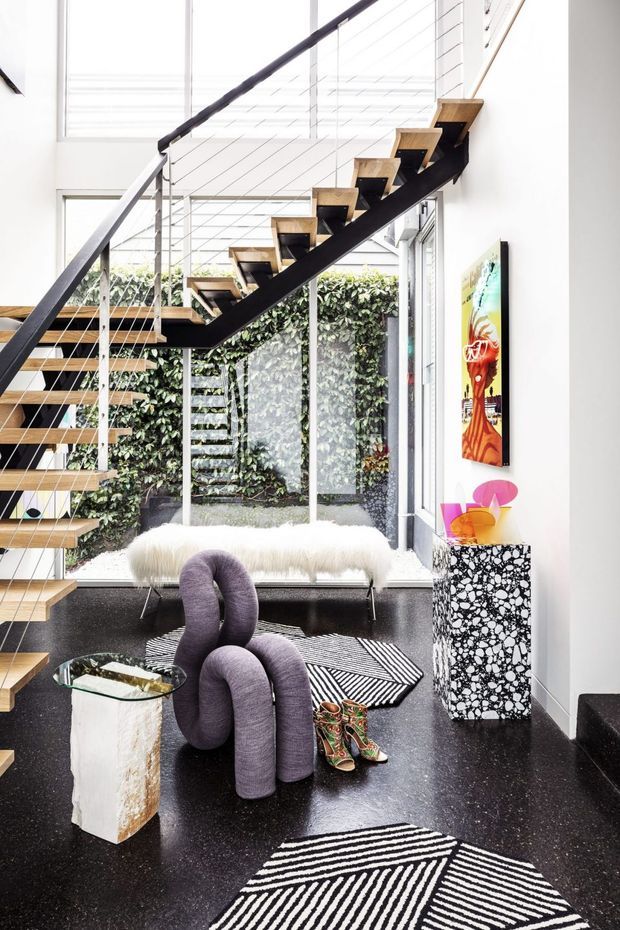 Bright, high-volume contemporary interior centred on a dramatic open-riser staircase with dark structural stringers, timber treads and fine cable balustrades. Full-height glazing overlooks a planted courtyard or living wall, while sculptural purple seating, a white fur bench, graphic black-and-white rugs, terrazzo-style side furniture and colourful artwork create a bold eclectic composition.