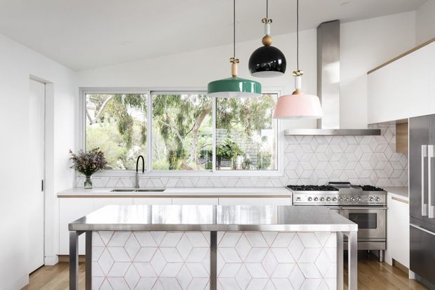 Bright contemporary kitchen with a large central island, white cabinetry, stainless-steel appliances, geometric patterned tile splashback, timber flooring, and three contrasting pendant lights. Large windows overlook leafy greenery.