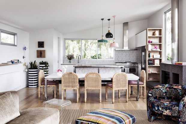 Bright, open-plan kitchen and dining area with a connected living space. The room features a white kitchen with marble-look patterned splashback, a large island with seating, timber flooring, rattan dining chairs, pendant lights, open shelving, and abundant natural light. A dark fireplace surround and patterned armchair add contrast.