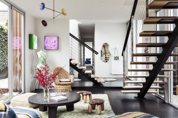 Bright, eclectic open-plan living space with a sculptural black staircase, open timber treads, glass balustrade, and a modernist mobile light fixture. Floor-to-ceiling glazing brings in natural light, while colourful artwork, a woven lounge chair, low round coffee table, stools, and layered rugs create a playful, curated interior.