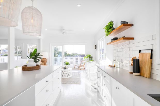 Bright white coastal-style kitchen with a large central island, white shaker cabinetry, pale grey benchtops, and a white horizontal tiled splashback. Open timber shelves with plants and cookware line the right wall, while woven pendant lights, indoor greenery, and a glazed dining or outdoor area beyond reinforce the airy, relaxed character.