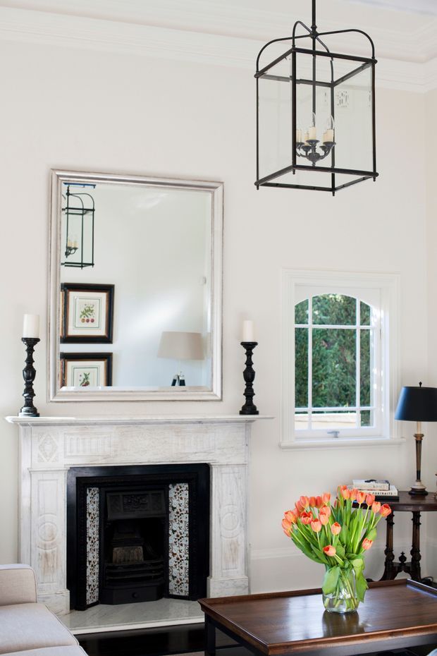 Bright, refined living room with a traditional fireplace and ornate white mantel, large framed mirror, sash window, dark wood coffee table, classic lamps, and a black metal glass lantern pendant. The room has pale walls, period detailing, and a restrained, elegant furnishing scheme.