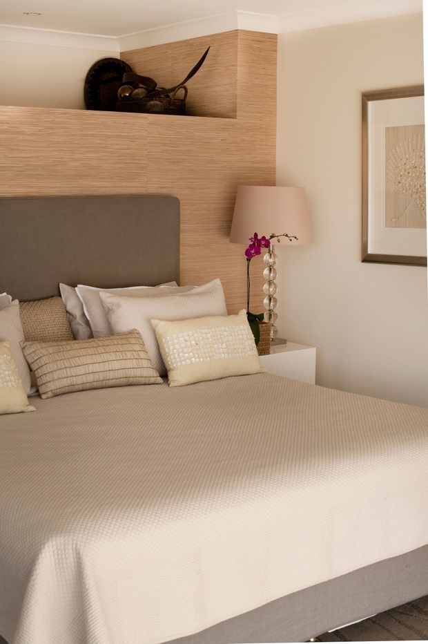 A warmly styled bedroom featuring a large upholstered bed with a grey headboard, layered cream and taupe bedding, and textured accent cushions. A timber-toned textured feature wall incorporates a recessed display shelf with sculptural decor. A glass-bead table lamp sits on a white bedside table, while framed artwork and neutral finishes complete the refined, softly lit interior.