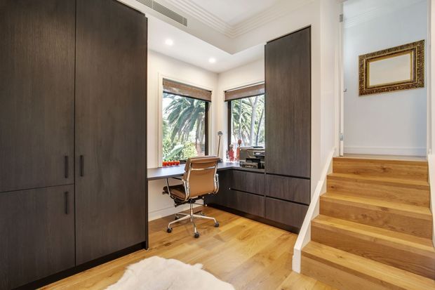Compact, refined home office with dark built-in cabinetry, a timber desk, and a wheeled leather office chair. Warm timber flooring continues up a short staircase, while large windows bring in abundant natural light and views of palm foliage. White walls, recessed downlights, detailed ceiling moulding, and a framed mirror or artwork complete the space.