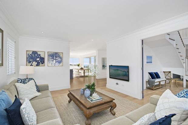 Bright, airy open-plan living room with neutral upholstered sofas, a substantial rustic timber coffee table, woven rug, coastal blue-and-white cushions, and floral artwork. A wall-mounted television sits beside an open circulation area leading to a dining space and large windows, while a staircase and small seating nook are visible to the right. White walls, decorative cornices, plantation shutters, and light timber flooring create a refined coastal-traditional interior.