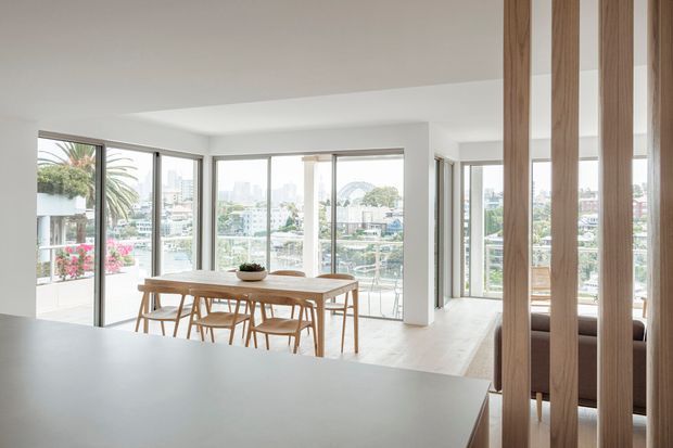 Bright contemporary open-plan interior with a dining area beside extensive corner glazing and a balcony, overlooking an urban waterfront or harbour setting. A light timber dining table and chairs sit on pale flooring, while vertical timber slats partially screen a lounge area. The space has a clean white ceiling, expansive windows, and abundant natural light.