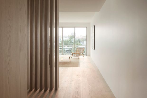 A minimalist, light-filled interior hallway or circulation space opening into a small sitting area. Pale timber flooring and a full-height timber slatted screen frame the left side, while smooth white walls and ceiling create a clean, quiet backdrop. At the far end, floor-to-ceiling glazing overlooks a leafy urban setting, with a simple timber lounge chair and low rug visible beyond.
