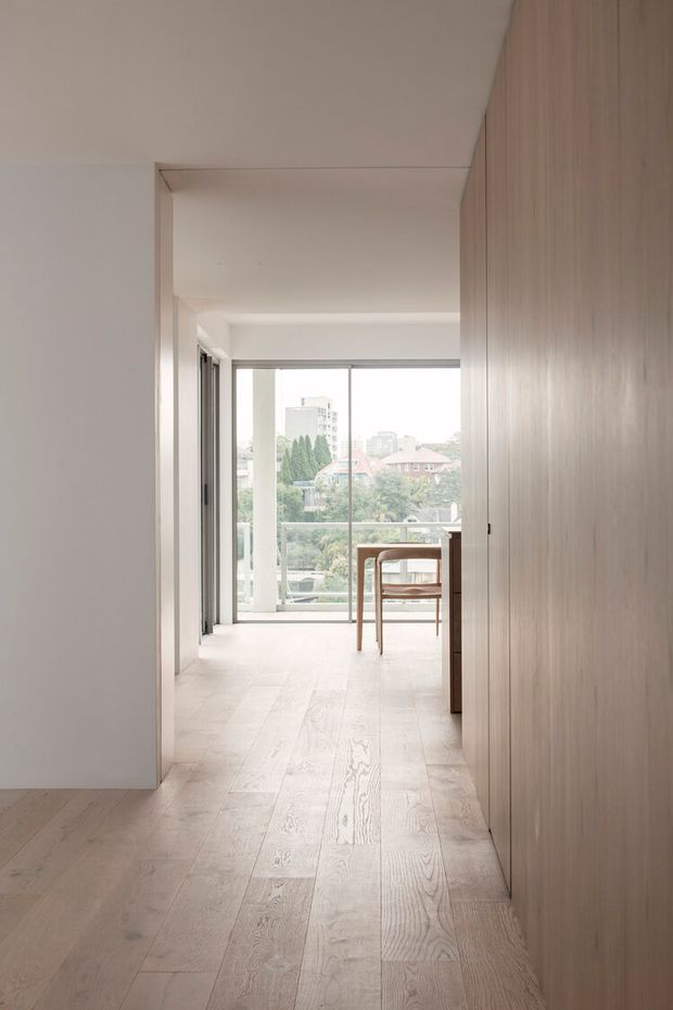 A minimalist residential interior viewed along a light timber-floored passage toward a bright dining area and full-height glazing. Pale built-in timber cabinetry lines the right side, while white walls and ceilings create a clean, understated composition. A small timber dining table and chairs sit near the windows, with an urban residential outlook beyond.