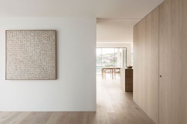 Bright, minimalist residential interior viewed from a hallway or entry area toward an open dining and living space. Pale timber flooring continues through the space, with a full-height light timber built-in wall of storage or cabinetry on the right and a white wall displaying framed artwork on the left. A dining table and chairs are visible near expansive glazing at the rear, creating an airy, calm atmosphere.