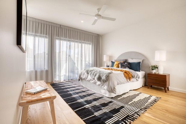 Bright contemporary bedroom with a large upholstered bed, layered neutral bedding, blue and mustard accents, sheer floor-to-ceiling curtains, light timber flooring, and a bold black-and-white patterned rug. A timber bedside table, bedside lamps, ceiling fan, and simple wall artwork complete the space.