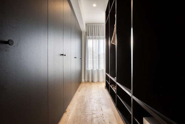 A narrow, well-organised walk-in wardrobe or dressing corridor with full-height dark built-in cabinetry on both sides, open shelving on the right, and a light timber floor leading to a softly curtained window. The space has a restrained contemporary, dark-and-light palette with recessed ceiling lighting.
