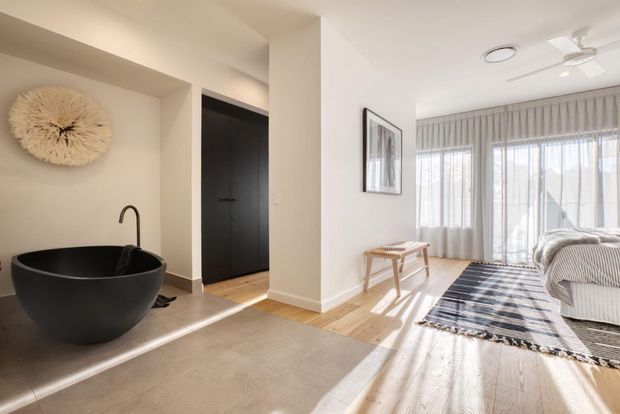Bright, open-plan residential interior showing a freestanding matte black bathtub in a recessed bathroom zone beside a dark doorway, opening into a sunlit living room. The living area has pale timber flooring, full-height sheer curtains, a striped sofa, a dark patterned rug, a small timber bench, and a white ceiling fan. Neutral plaster walls and abundant natural light create a contemporary, calm atmosphere.