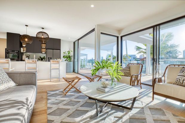 Bright open-plan living room with a contemporary kitchen behind it, featuring expansive floor-to-ceiling glazing, a pale round coffee table, upholstered seating, patterned rug, timber flooring, and woven pendant lights. The glazing opens to a covered terrace with coastal or waterfront views and palms visible outside.