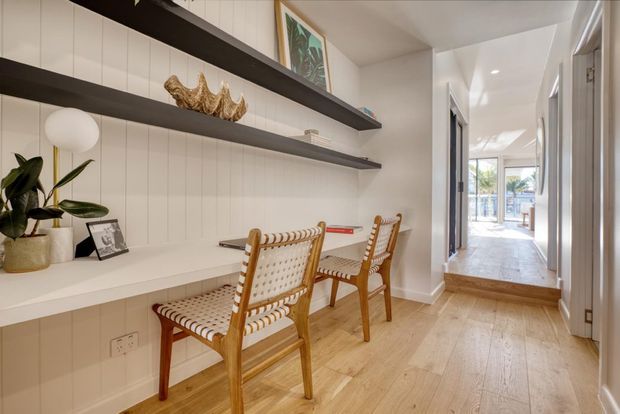 Bright, narrow built-in workspace or study nook with a long white desk, two woven timber-framed chairs, vertical wall panelling, dark floating display shelves, and light timber flooring. A hallway leads toward a glazed outdoor area, creating an airy, contemporary coastal feel.