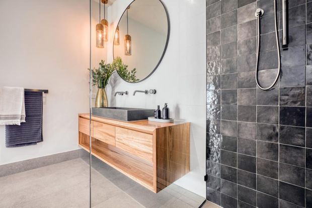 Contemporary bathroom featuring a floating timber vanity with a dark rectangular basin, wall-mounted tapware, a large round mirror, pendant lights, and a walk-in shower lined with dark grey tiles behind clear glass.
