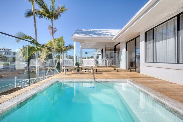 Bright exterior view of a modern single-storey residence with a turquoise swimming pool in the foreground, timber decking, glass pool fencing, palm trees, and a covered outdoor entertaining terrace beside full-height glazing. The setting appears warm and coastal, with neighbouring waterfront-style homes visible beyond the pool fence.