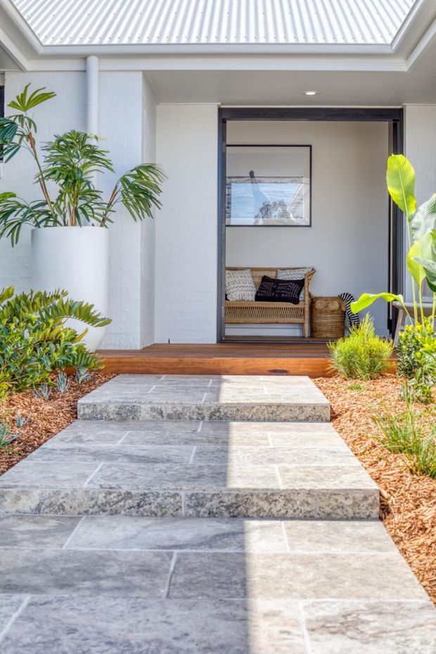 Bright exterior entry courtyard leading up broad stone steps to a contemporary home's open doorway. The facade is painted white with a corrugated metal roof, dark-framed glazing, timber threshold decking, tropical potted plants, and landscaped garden beds. A woven bench is visible inside the entry.
