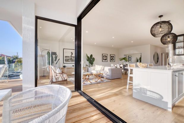 Bright open-plan coastal-style interior viewed from a balcony or terrace, with timber decking in the foreground leading through large black-framed sliding glass doors into a living, dining and kitchen area. The room features pale timber flooring, white cabinetry and walls, a white sectional sofa, woven furniture, indoor plants, framed artwork and distinctive black woven pendant lights. Strong indoor-outdoor connection and abundant natural light define the space.