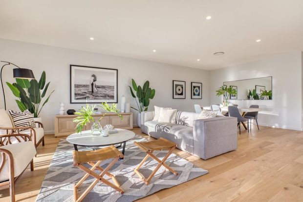 Bright, airy open-plan living room with light timber flooring, white walls, a large grey sectional sofa, patterned rug, round coffee table, leather-and-wood lounge chairs, indoor plants, framed artwork, and a dining area visible at the rear. A large wall mirror reflects the room and dining space. Recessed ceiling lighting and restrained contemporary furnishings create a clean, inviting atmosphere.