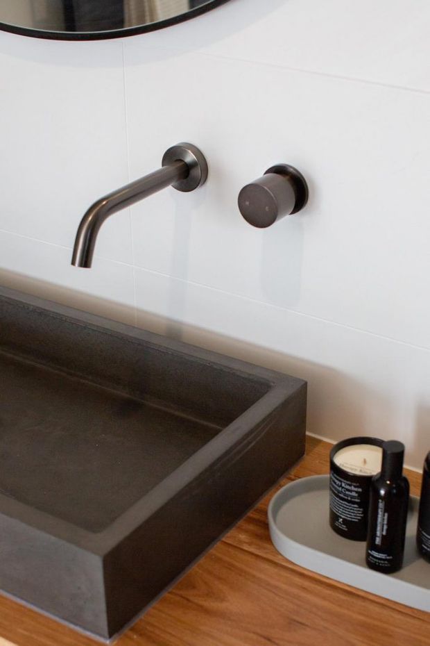 Close-up of a contemporary bathroom vanity featuring a wall-mounted brushed dark-metal basin tap and separate cylindrical control on a white tiled wall. A large rectangular dark stone or concrete vessel basin sits on a warm timber vanity surface, with toiletries in a tray beside it and part of a round mirror visible above.