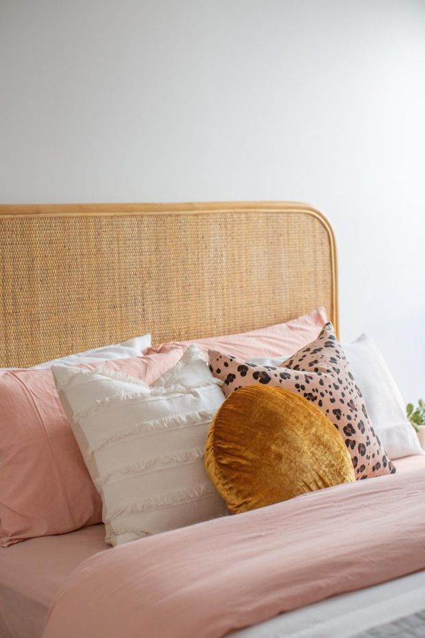 Close-up of a softly styled bed with a rounded woven rattan/cane headboard, blush pink bedding, white fringed pillows, a leopard-print cushion and a mustard-gold round cushion. The bright neutral wall and warm natural textures create a playful, cozy bedroom setting.
