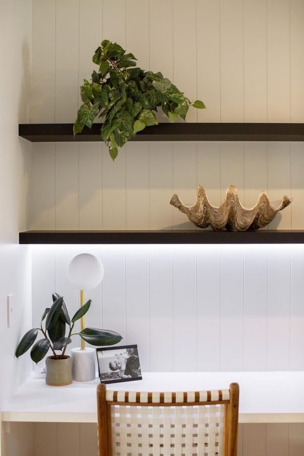 A close-up interior view of a compact home office or study nook with a white vertical-panel wall, two dark floating shelves, integrated under-shelf lighting, potted plants, sculptural shell decor, and a small desk with a woven timber chair. The styling is warm, refined, and contemporary with natural accents.