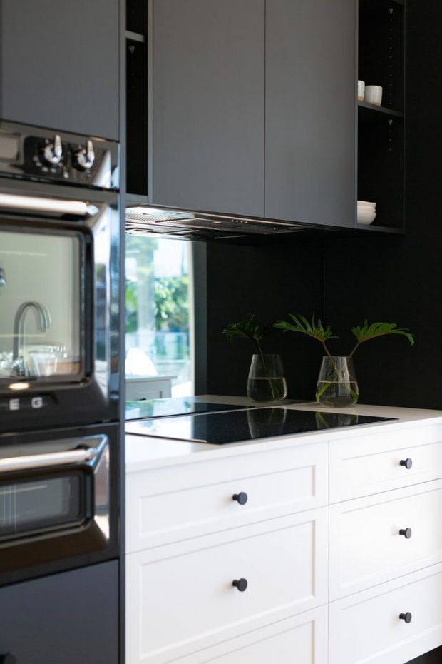 A compact, refined kitchen with white shaker-style base cabinetry and dark grey upper cabinetry, a glossy black benchtop and black splashback, integrated stainless-steel ovens, and open black shelving with ceramic vessels. Simple greenery in glass vases adds a natural touch, while a window brings daylight into the space.