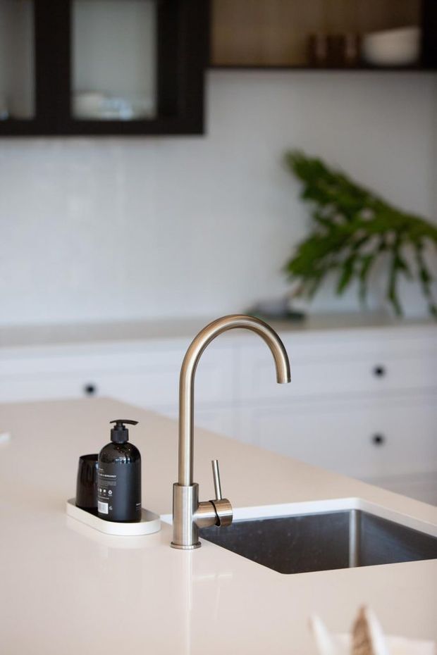 Close-up view of a contemporary kitchen sink and curved brushed-metal tap set into a pale engineered-stone benchtop. White cabinetry with dark round pulls and a dark-framed window or shelving element forms the background, with a small plant and soap dispenser adding detail.