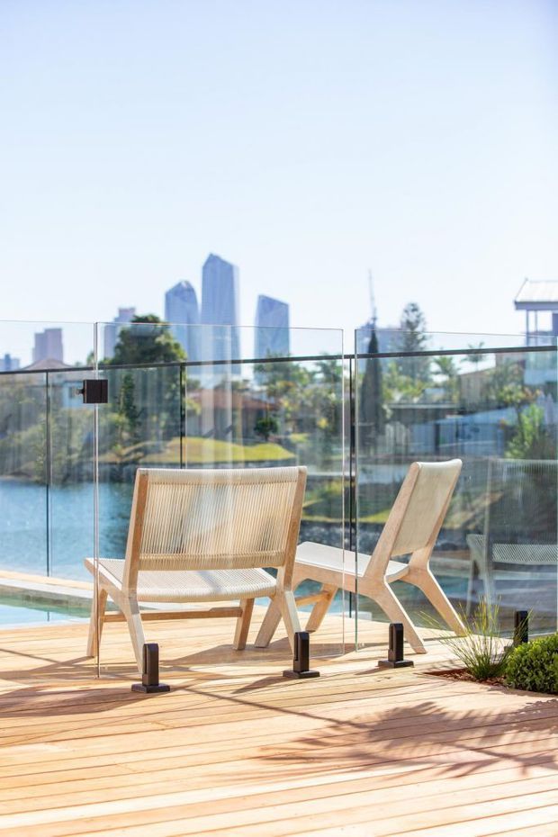 A sunlit outdoor timber deck with two light timber lounge chairs positioned beside a clear glass balustrade. Water is visible beyond the deck, with landscaped greenery and a modern city skyline in the background. The scene has a relaxed, contemporary waterfront terrace character.