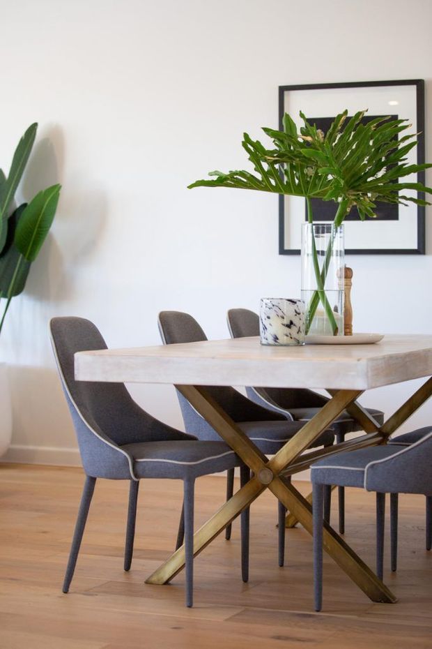 A bright, refined dining room featuring a substantial light timber dining table with crossed trestle-style legs, upholstered charcoal-grey dining chairs, pale timber flooring, white walls, large indoor plants, and a black-framed artwork. A vase of tropical foliage forms the table centrepiece.