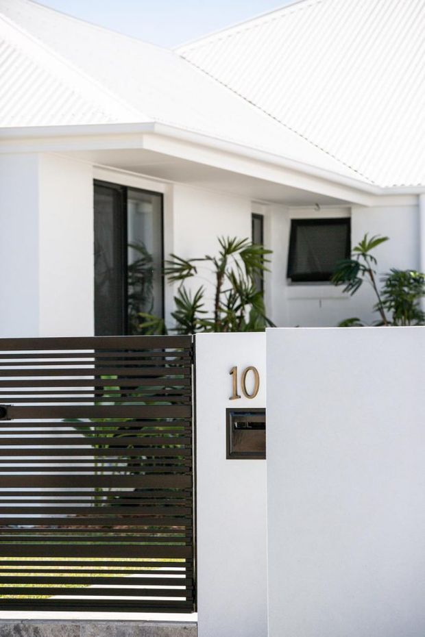 Front exterior view of a contemporary white-rendered residence with a bright corrugated metal pitched roof, covered entry porch, dark horizontal-slat gate, white boundary wall, house number, letterbox, dark-framed glazing, and tropical plants in the entry courtyard.