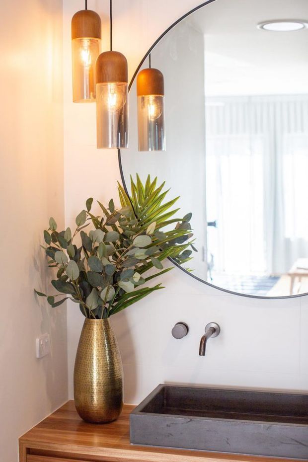 A refined bathroom vanity vignette featuring a rectangular dark stone basin set into a timber vanity, wall-mounted brushed metal tapware, a large round mirror, and three warm amber and clear pendant lights. A tall hammered brass vase with leafy greenery adds an organic decorative accent. The palette is warm timber, white walls, dark grey stone, brass, and amber glass.