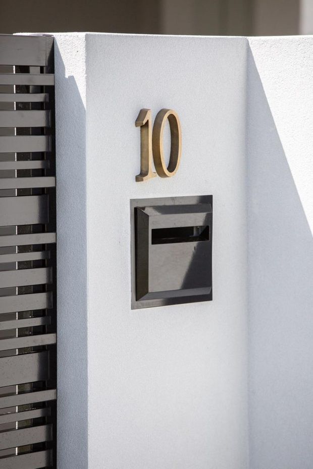 Close-up exterior detail of a white textured rendered wall featuring raised metallic-gold house numbers "10" above a recessed dark metal letterbox. A black horizontal slatted gate or screen is visible along the left edge, with strong sunlight creating crisp shadows.