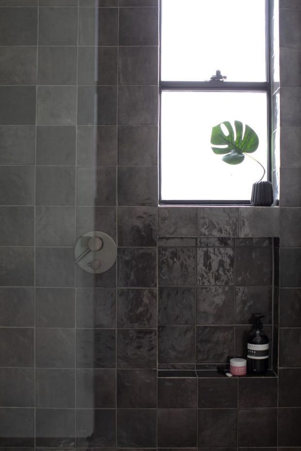 A moody tiled bathroom or shower area with dark grey ceramic wall tiles, a recessed shelf holding toiletries, a circular wall control, and a black-framed frosted or bright window with a small potted leaf plant.