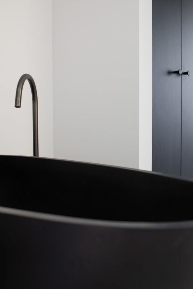 Close-up architectural photograph of a minimalist bathroom featuring a large matte black freestanding bathtub in the foreground, a dark curved faucet, pale grey walls, and tall dark cabinetry with simple black hardware.