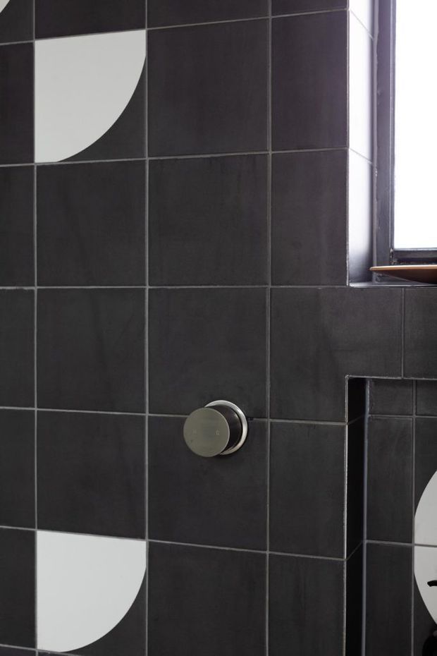Close-up of a dark tiled bathroom wall featuring a round brushed-metal control, a narrow window, and bold white quarter-circle geometric tile accents. The dark grout and monochrome pattern create a graphic, contemporary look.