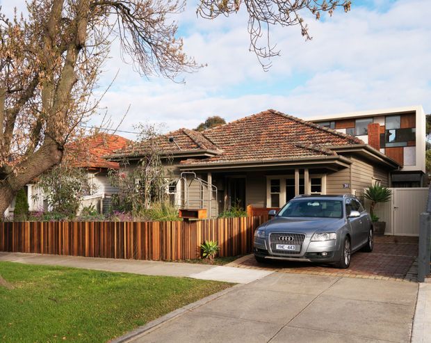 Street-facing view of a traditional single-storey timber-clad bungalow with a terracotta tiled hipped roof, front veranda, timber fencing and driveway. A contemporary multi-storey extension is visible behind and to the right, creating a clear contrast between the older dwelling and newer addition. Mature trees and lawn frame the suburban setting.