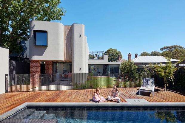 Contemporary residential exterior viewed from a pool and expansive timber deck. The home features rounded rendered forms, exposed red-brick walls, dark metal fencing, large windows, and a flat-roofed upper level. A neighbouring pitched-roof dwelling and mature trees are visible in the background.