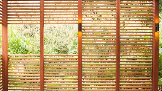 A close-up exterior view of a timber louvred screen or privacy wall, composed of warm reddish-brown vertical posts and closely spaced horizontal slats. The open slats allow filtered views of dense tropical planting beyond, while small integrated wall lights are mounted between the screen sections.